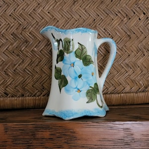 May include: A small white ceramic pitcher with a blue rim and handle. The pitcher is decorated with hand-painted blue flowers and green leaves.