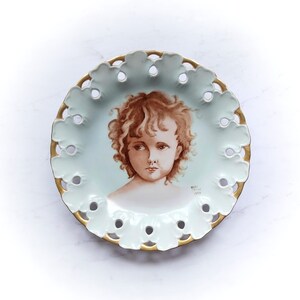 May include: A white porcelain plate with a gold rim and a cutout design. The plate features a painted portrait of a young girl with brown hair and brown eyes. The text "Mary Italia 1972" is printed on the plate.