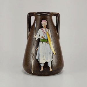 May include: Brown ceramic vase with two handles, featuring a painted figure of a man in traditional attire. The figure holds a spear and a shield. The vase has a textured surface and a small opening at the top.