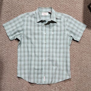 May include: A short-sleeved, button-down shirt in a plaid pattern of light green, white, and dark green. The shirt has a collar and a small penguin logo on the chest. The brand name is visible on the collar.