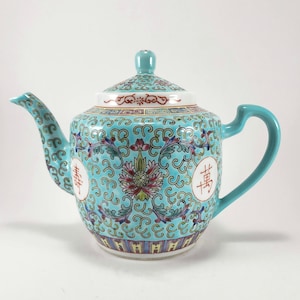 May include: A turquoise porcelain teapot featuring a detailed floral scroll pattern in pink, green, and blue. The body displays two white circular medallions with red Chinese characters. It includes a matching lid with a knob and a curved handle.