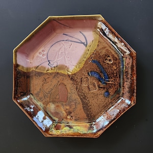 May include: An octagonal ceramic plate featuring a dual-tone glaze of dusty pink and deep brown. The surface displays abstract dark blue brushstrokes, incised spiral patterns, and small circular indentations, with a glossy, textured finish.