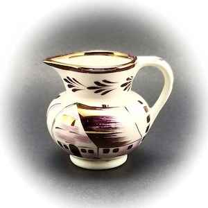 May include: A small white ceramic creamer with a purple and gold painted design of a house. The creamer has a handle and a gold rim.