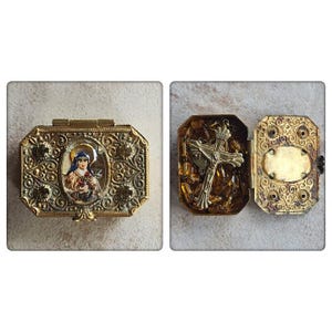 May include: A rectangular gold-toned metal hinged box with an ornate filigree pattern and a central oval inlay depicting a figure in a habit holding roses. The interior contains a silver-toned crucifix and amber-colored beads for a rosary.