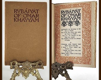 Pode incluir: Um livro vintage intitulado "Rubaiyat of Omar Khayyam" é exibido aberto e fechado em suportes decorativos de latão. A capa é marrom claro com texto marrom. A página aberta tem bordas e texto ornamentados.