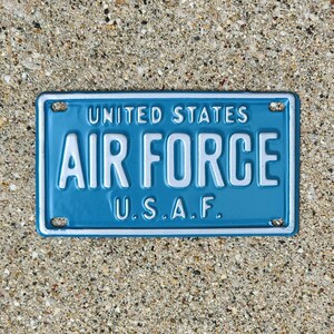 May include: A rectangular, light blue metal sign with the words "UNITED STATES AIR FORCE" in white block letters. Below, the letters "U.S.A.F." are also in white. The sign has a white border and four mounting holes.