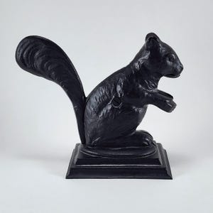 May include: A black cast iron nutcracker shaped like a squirrel. The squirrel is depicted in a seated position with its tail arched upward, mounted on a square base. The metal surface has a textured finish, and a visible screw is on the side.
