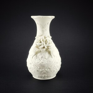 May include: White ceramic vase with an ornate design and a floral accent. The vase has a pearlescent finish and a delicate floral design.