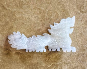 May include: A carved, white dragon figurine with intricate details. The mythical creature is depicted in a side profile, with a spiky mane and tail. The figurine is made of a translucent material.