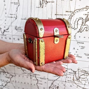 Puede incluir: Un pequeño cofre de madera rojo con detalles metálicos dorados. El cofre se sostiene en manos abiertas sobre un fondo de mapa antiguo. El cofre tiene una parte superior curvada y un cierre de metal.