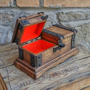 May include: A small, wooden treasure chest with a hinged lid, open to reveal a bright red interior. The chest is accented with black metal hardware and sits on a wooden base. The wood has a distressed finish.
