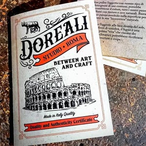 May include: A white card with black and orange text and a black and white illustration of the Colosseum. The text reads "DOREALI STUDIO ROMA BETWEEN ART AND CRAFT Made in Italy Quality Quality and Authenticity Certificate".