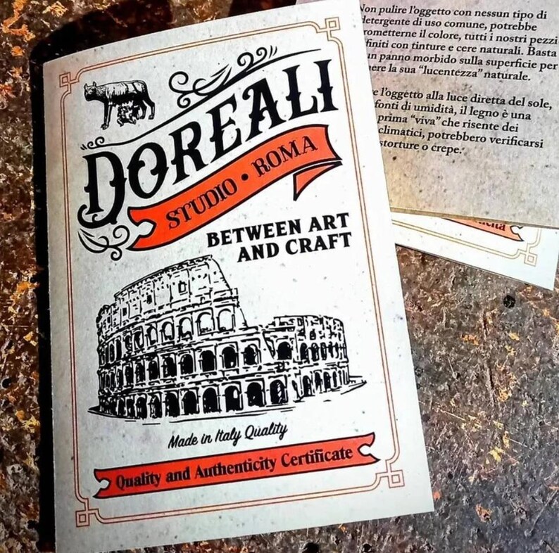 May include: A white card with black and orange text and a black and white illustration of the Colosseum. The text reads "DOREALI STUDIO ROMA BETWEEN ART AND CRAFT Made in Italy Quality Quality and Authenticity Certificate".