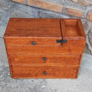 May include: A wooden box with three drawers and a hinged top. The box is stained a rich brown color and has black metal hinges and handles.