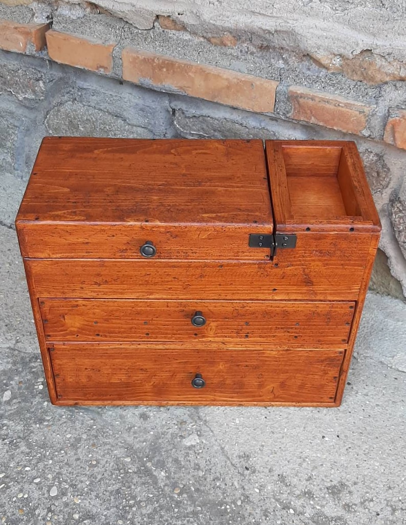 May include: A wooden box with three drawers and a hinged top. The box is stained a rich brown color and has black metal hinges and handles.