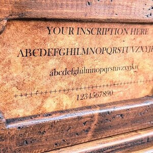 May include: A wooden box with a hinged lid and a small plaque on the front. The plaque has a brown background and the text "YOUR INSCRIPTION HERE" in black lettering. Below the text is a line of the alphabet in uppercase and lowercase letters, followed by a row of numbers from 1 to 9.