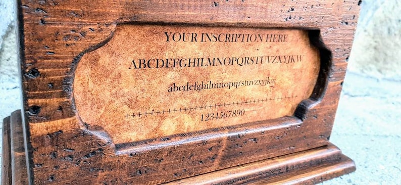 May include: A wooden box with a hinged lid and a small plaque on the front. The plaque has a brown background and the text "YOUR INSCRIPTION HERE" in black lettering. Below the text is a line of the alphabet in uppercase and lowercase letters, followed by a row of numbers from 1 to 9.
