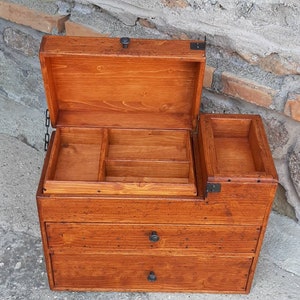 May include: A wooden box with a hinged lid and two drawers. The box has a brown stain and black metal hardware. The lid is open and reveals a small compartment with dividers.