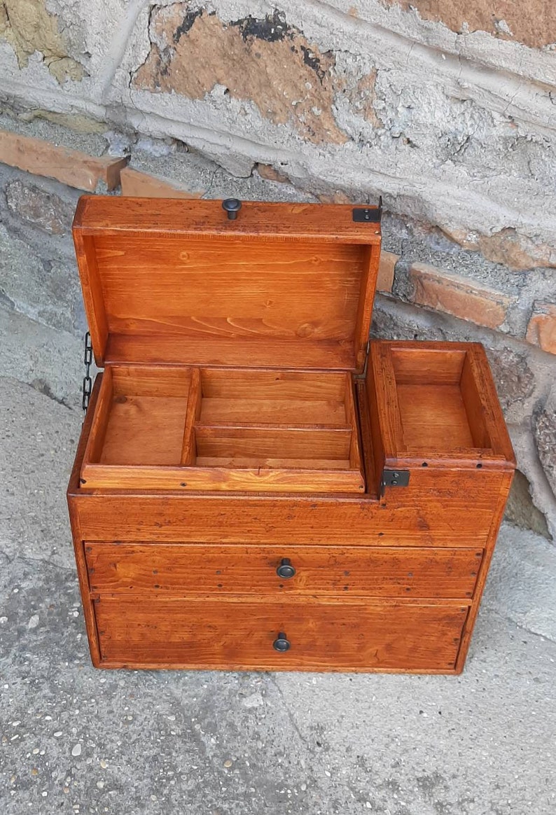 May include: A wooden box with a hinged lid and two drawers. The box has a brown stain and black metal hardware. The lid is open and reveals a small compartment with dividers.