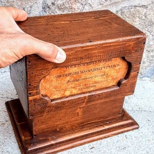 May include: A wooden box with a hinged lid and a small base. The box has a label on the front that says "YOUR INSCRIPTION HERE" and includes the alphabet and numbers. The box is brown and has a rustic appearance.