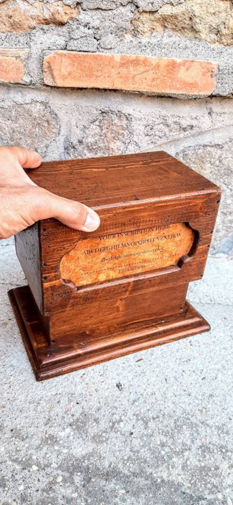 May include: A wooden box with a hinged lid and a small base. The box has a label on the front that says "YOUR INSCRIPTION HERE" and includes the alphabet and numbers. The box is brown and has a rustic appearance.