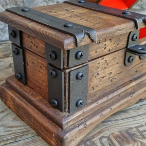 May include: A small, wooden treasure chest with a hinged lid, featuring dark metal accents and screws. The chest has a distressed, rustic appearance with visible wood grain and a reddish-orange interior. The chest sits on a wooden base.