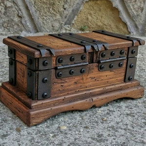 May include: A small, wooden treasure chest with a distressed finish. The chest is rectangular with a hinged lid and is reinforced with metal bands and rivets. The wood has a warm, brown tone, and the metal accents are dark. The chest sits on a matching base.