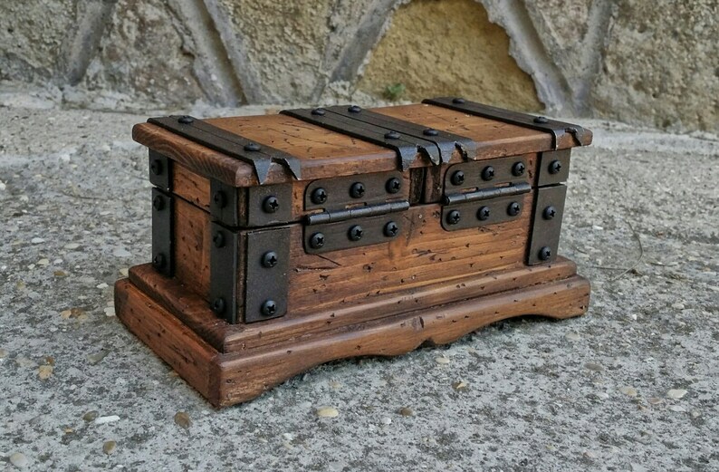 May include: A small, wooden treasure chest with a distressed finish. The chest is rectangular with a hinged lid and is reinforced with metal bands and rivets. The wood has a warm, brown tone, and the metal accents are dark. The chest sits on a matching base.