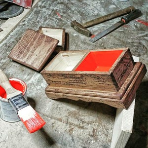 May include: A wooden box with a hinged lid, stained in shades of brown, with a bright red interior compartment. A paintbrush dipped in red paint and a can of paint are in the foreground. Tools like a hammer and file are in the background, suggesting a woodworking project.