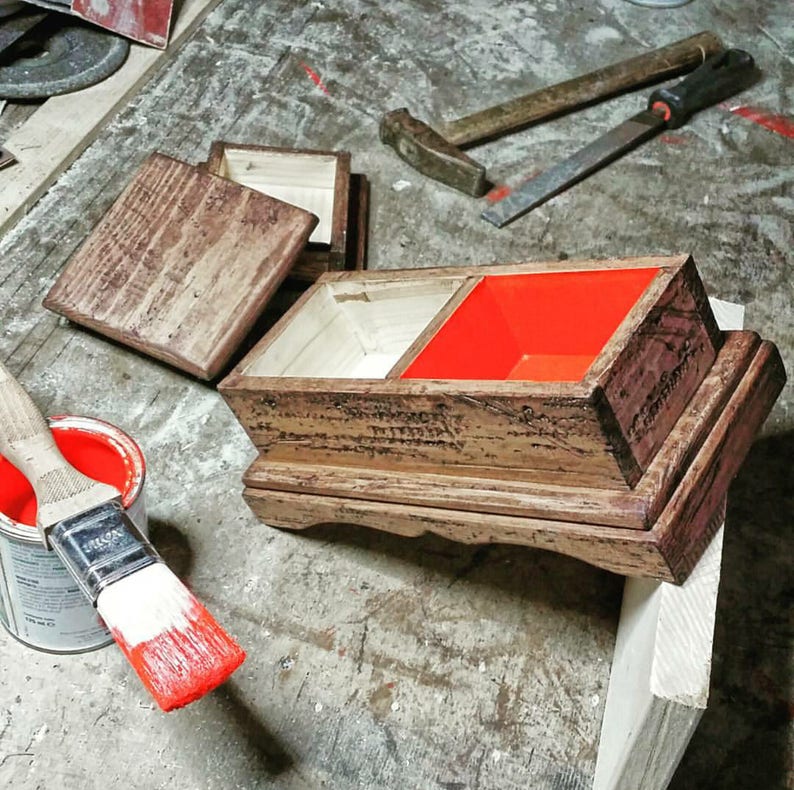 May include: A wooden box with a hinged lid, stained in shades of brown, with a bright red interior compartment. A paintbrush dipped in red paint and a can of paint are in the foreground. Tools like a hammer and file are in the background, suggesting a woodworking project.