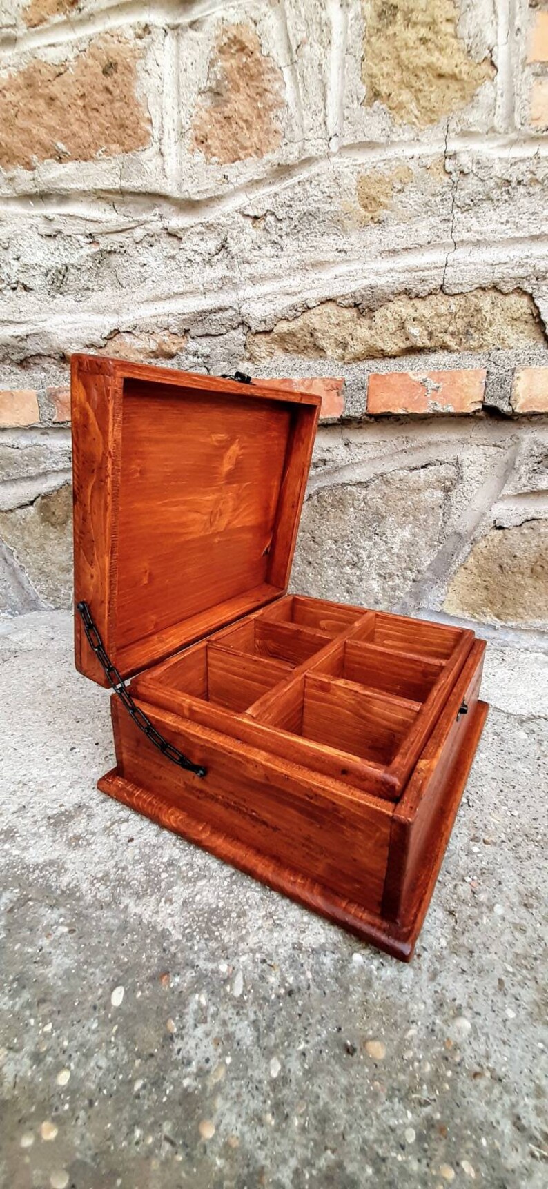 May include: A wooden box with a hinged lid and a metal chain closure. The box has six compartments inside and is stained a rich brown colour.