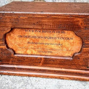 May include: A wooden box with a hinged lid and a label on the front. The label has the text "YOUR INSCRIPTION HERE" and the alphabet in a decorative font. The box is brown and has a rustic appearance.