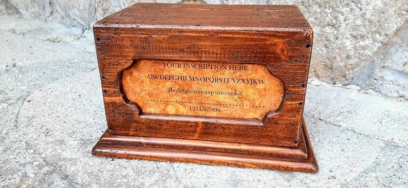 May include: A wooden box with a hinged lid and a label on the front. The label has the text "YOUR INSCRIPTION HERE" and the alphabet in a decorative font. The box is brown and has a rustic appearance.