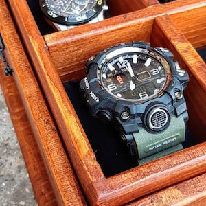 May include: A wooden watch box with a hinged lid, featuring multiple compartments for storing watches. The box is made of rich, reddish-brown wood with a smooth finish. Three watches are visible, including a black and silver chronograph watch, a silver watch with a white face, and a black and green digital watch with the text "WATER RESIST" on the band.