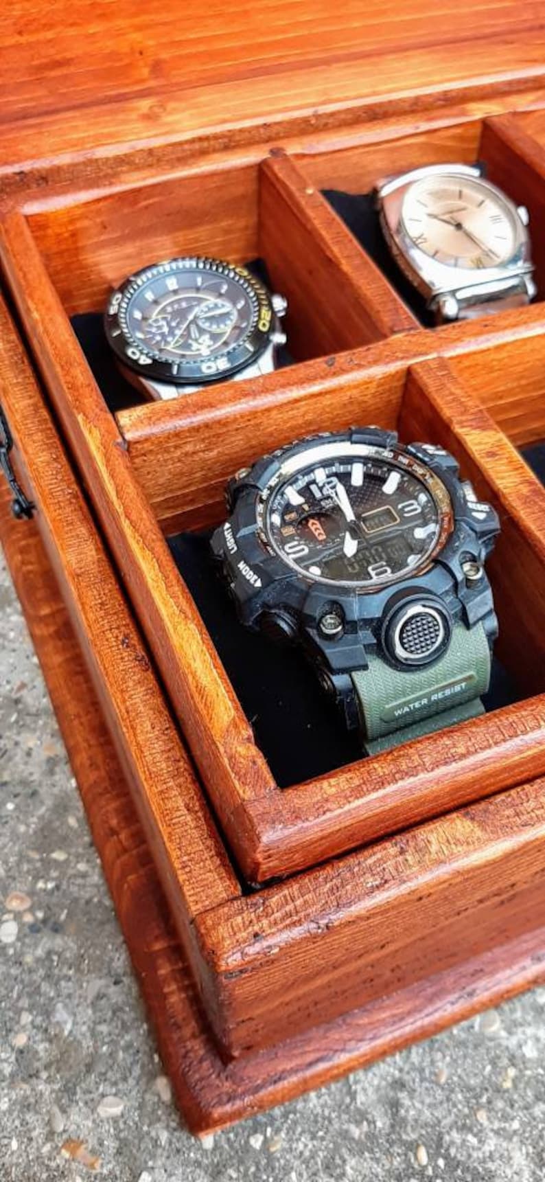 May include: A wooden watch box with a hinged lid, featuring multiple compartments for storing watches. The box is made of rich, reddish-brown wood with a smooth finish. Three watches are visible, including a black and silver chronograph watch, a silver watch with a white face, and a black and green digital watch with the text "WATER RESIST" on the band.