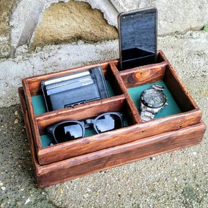 May include: A wooden organizer with multiple compartments for storing items like a phone, wallet, sunglasses, and a watch. The organizer has a dark brown stain and a green felt lining.