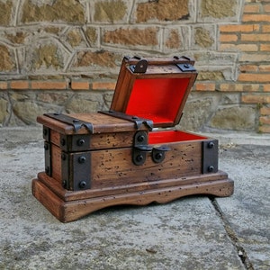 May include: A small, wooden treasure chest with a hinged lid, open to reveal a red interior. The chest is brown with metal accents and sits on a matching base. The chest is designed to look antique.