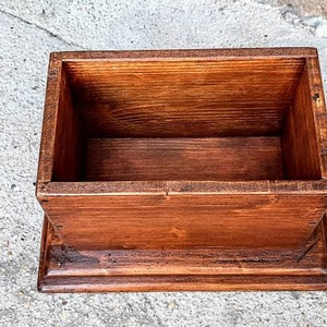 May include: A wooden box with a hinged lid and a base. The box is made of natural wood with a rich brown stain. The box is empty and ready to be filled.