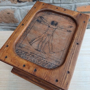 May include: A wooden box with a hinged lid, stained in a warm brown hue. The lid features an engraved image of Leonardo da Vinci's Vitruvian Man. The box is constructed with visible joinery and small, dark-colored nail heads.