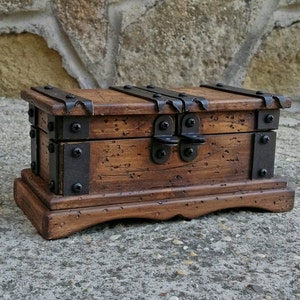 May include: A small, wooden treasure chest with a dark brown finish. The chest features metal bands and hinges, and a clasp closure. The chest sits on a decorative base, and is designed to look antique.