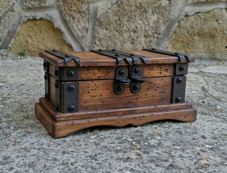 May include: A small, wooden treasure chest with a dark brown finish. The chest features metal bands and hinges, and a clasp closure. The chest sits on a decorative base, and is designed to look antique.