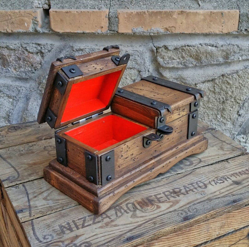 May include: A small, wooden treasure chest with a hinged lid, open to reveal a bright red interior. The chest is accented with black metal hardware and sits on a wooden base. The wood has a distressed finish.