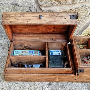 Puede incluir: Una caja de madera con tapa abatible, dividida en compartimentos. La caja contiene gafas de sol, tarjetas y llaves. La caja está hecha de madera natural con un acabado rústico.