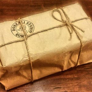 May include: A rectangular package wrapped in tan paper and tied with twine. The package has a circular stamp that reads "Doreali Studio 100% Hand Made in Italy Roma". The twine is tied in a bow on top of the package.