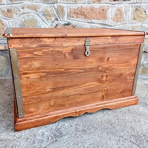 May include: A wooden storage chest with a hinged lid and brass hardware. The chest is stained a rich brown color and has a decorative trim around the edge.