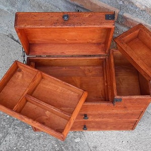 May include: A wooden box with a hinged lid and three drawers. The box is made of light brown wood and has black metal accents. The box is open and the drawers are pulled out.