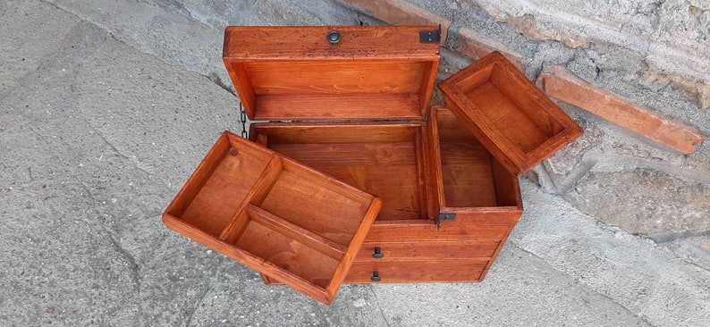 May include: A wooden box with a hinged lid and three drawers. The box is made of light brown wood and has black metal accents. The box is open and the drawers are pulled out.