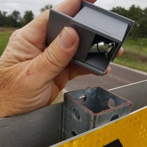 May include: A gray 3D printed box with a silver metal object inside. The box is being held by a hand and is being placed on a metal post with holes. The post is attached to a yellow and black sign.