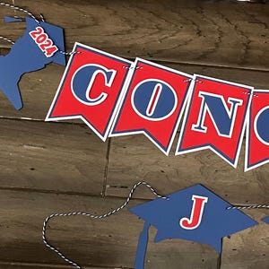 May include: A graduation banner with the word "CONGRATS" in red and blue pennants, and a separate banner spelling "JON" in blue graduation caps. The year "2024" is on the ends of the "CONGRATS" banner.