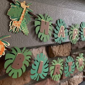 Puede incluir: Una guirnalda de cumplea&ntilde;os con tem&aacute;tica de jungla con las palabras "HAPPY BIRTHDAY" escritas en letras marrones sobre recortes de hojas verdes. La guirnalda incluye una jirafa y un le&oacute;n. La guirnalda est&aacute; ensartada en una cinta marr&oacute;n.
